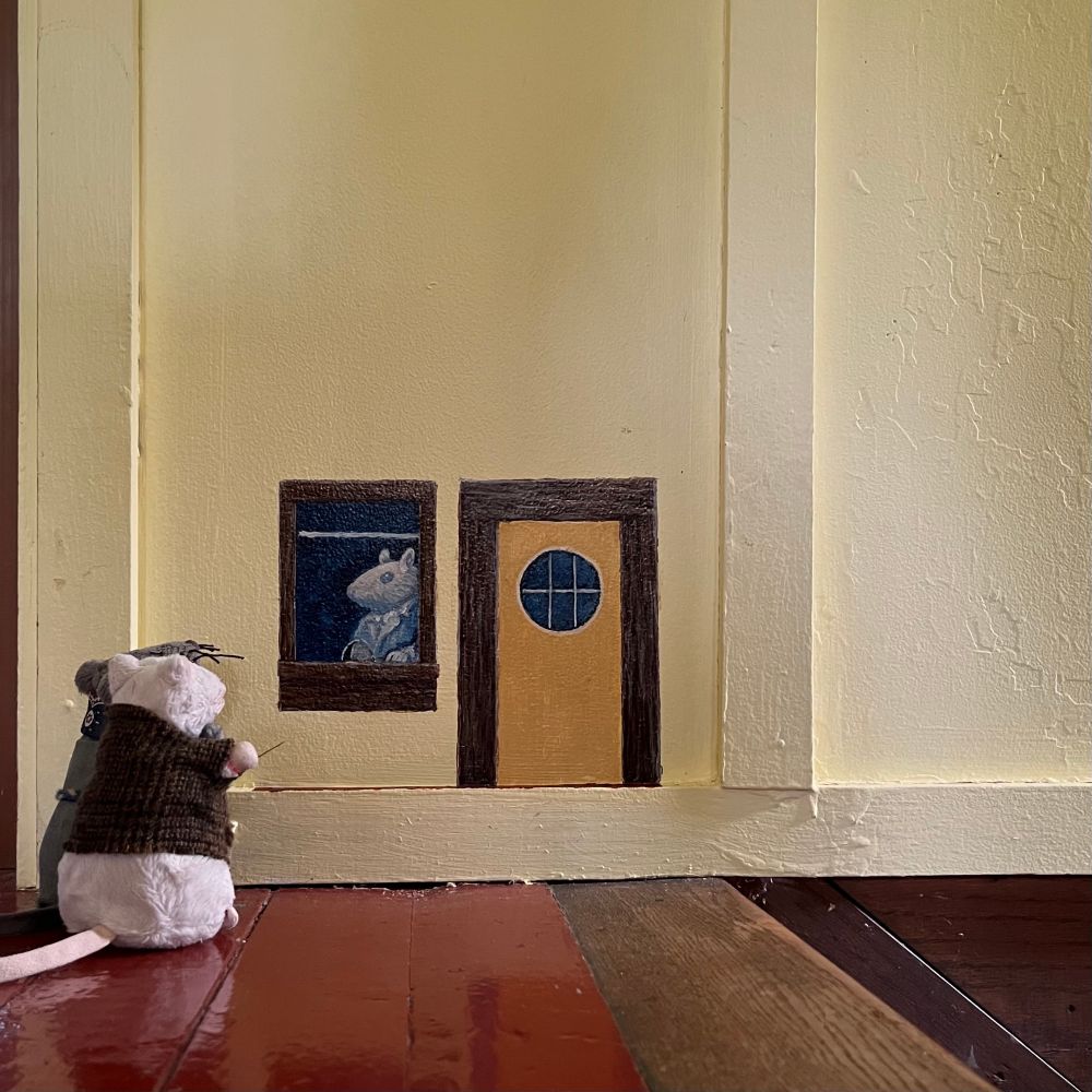 Two plush mice at the top of a staircase looking at an unfinished painting of a mouse-sized ticket booth and door for the planned cottage-wide metro system. 