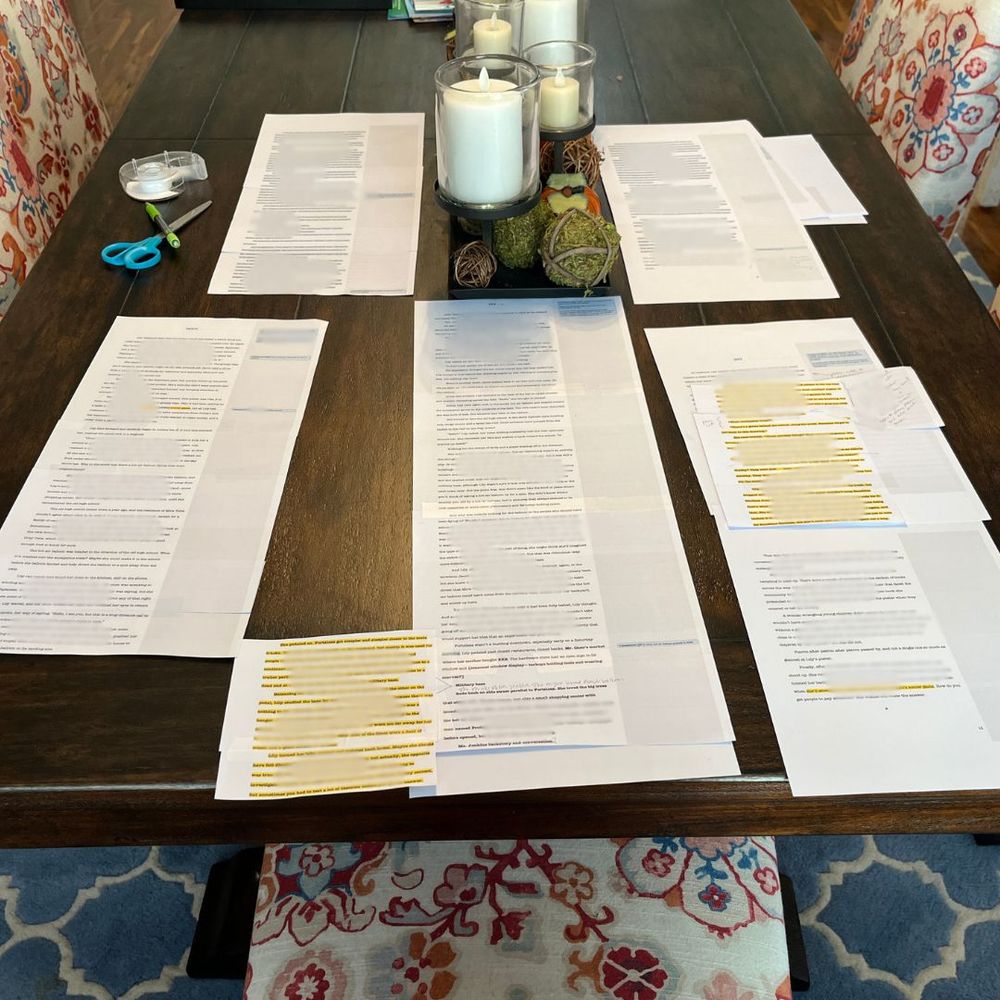 printed pages of a manuscript on a dining table. Pages have highlighted sections and have been cut up and taped back together to form long chains.