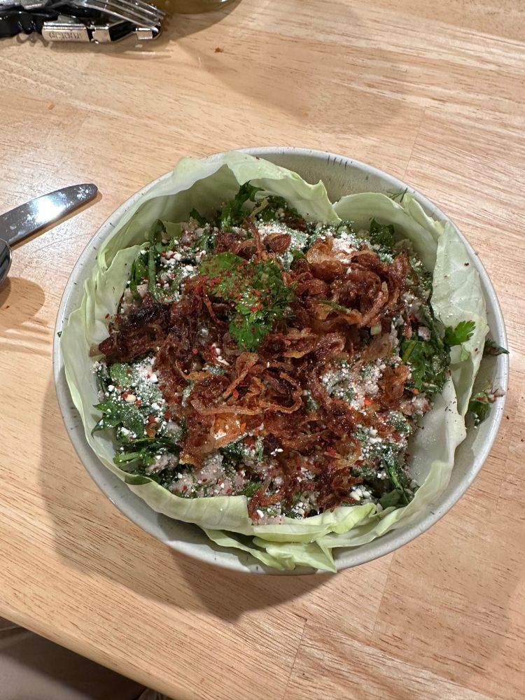 A bowl lined with cabbage leaves and filled with Larb: ground pork, dill, cilantro, mint, Thai basil, lime juice, fish sauce, fresh and dried Thai chilis, toasted ground rice, and shallots. 