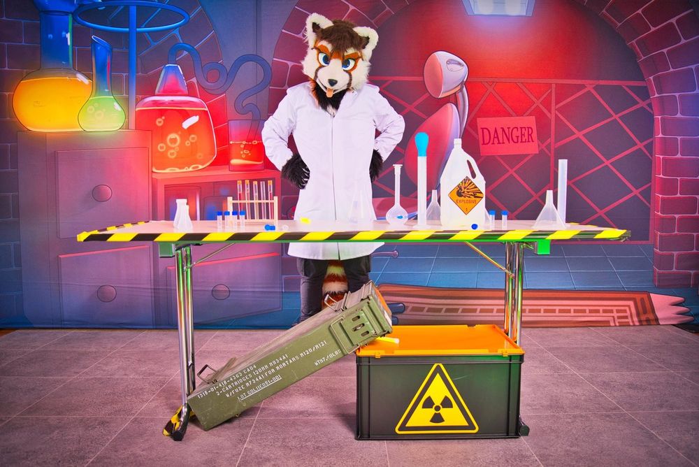 Me in my red panda fursuit wearing a lab coat. In front of me is a large desk, on which various fake chemical equipment sits as props.
