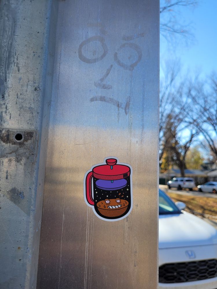 A sticker of a coffee press. The coffee grounds at the bottom have a face on them which is grinning with delight.