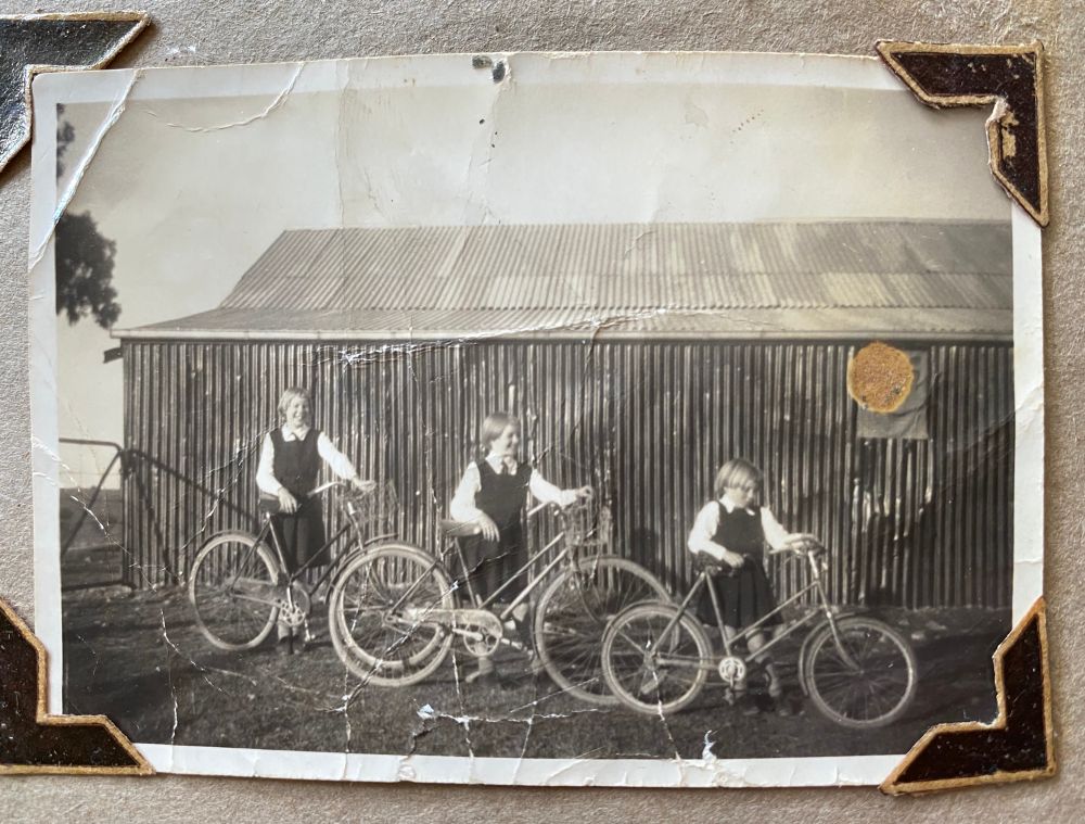 Black and white photo of three white girls aged about 13, 9 and 5, with blonde bowl cuts, in matching dark Barfold State School square-necked tunics and ties with white shirts underneath. c1950, maybe. Each is holding a bicycle. They're standing in order of birth left to right, near a farm gate and a shed made entirely of corrugated iron. Wanda and Linda are smiling. Glenda is looking at her handlebars. The photo is shown with arrow shaped corners, in an old photo album. 