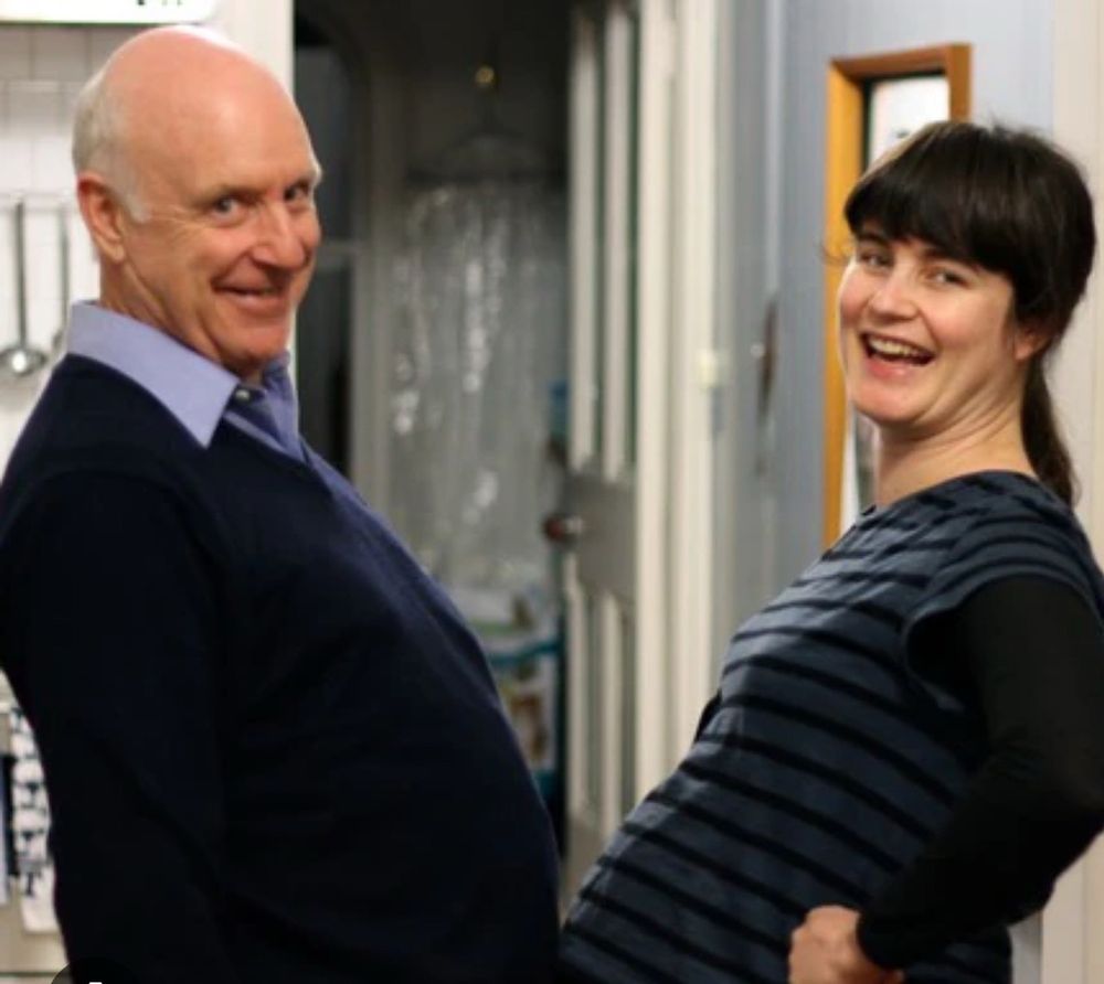 Satirist John Clarke in a jumper opposite his daughter, writer & director Lorin Clarke. They are grinning at the camera in characteristic ways. Both have significant profile tums; Lorin because she was up the duff at the time. 