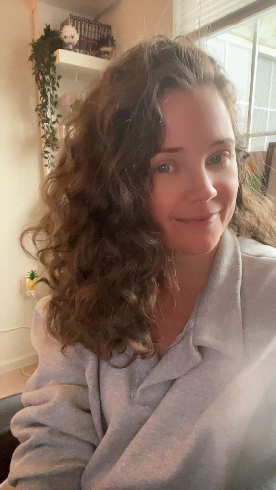 This is a picture of me (mostly my hair, which is brown and right now features a lot of shiny, beautiful curls). I’m in a grey sweatshirt and sitting in front of a window.