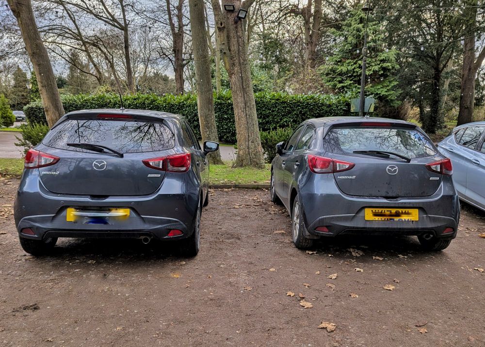Two dark grey Mazda 2 cars parked side by side, seen from the rear. 