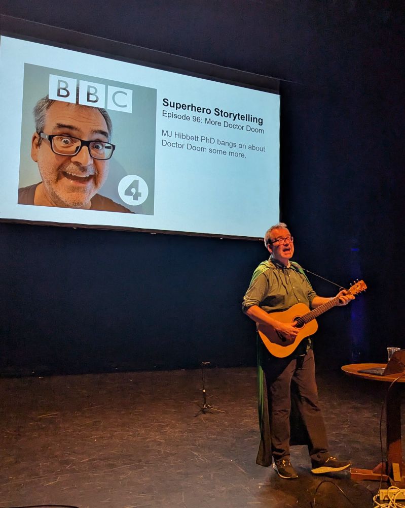 MJ Hibbett performing on-stage playing guitar and wearing a cape. Behind him on a screen is a slide promoting his fictitious (so far) Radio 4 series on superheroes. 