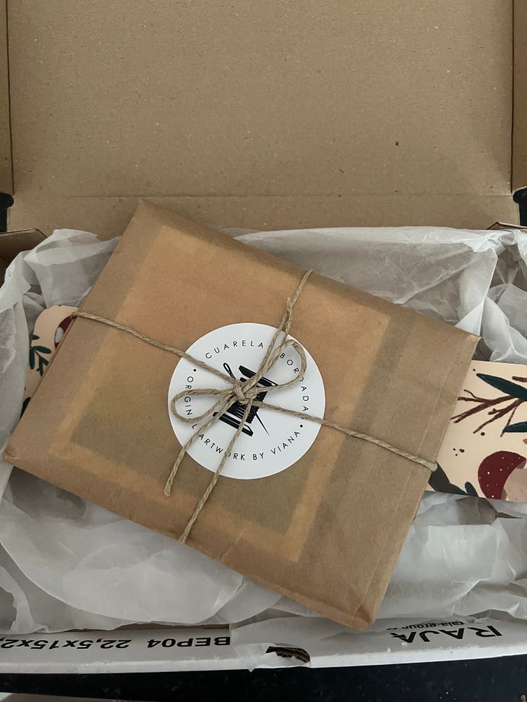 Imagen de un paquete a medio abrir, se ve algo envuelto en papel reciclado marrón, atado con cordel y una pegatina redonda con la imagen de un carrete de hilo con una aguja enganchada y texto que dice “acuarela bordada, original artwork by Viana” todo sobre una caja de cartón que también contiene papel de envolver blanco y un marcapáginas ilustrado.  