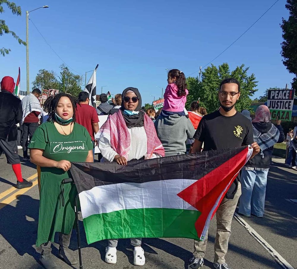 Three members of the GEOC (Wayne State's Graduate Employees Organizing Committee) steering committee stand as part of the 2025 Labor Day Parade in Detroit, Michigan, holding a Palestinian flag. Behind them, other comrades display flags, keffiyehs, and posters promoting peace and justice for Gaza.