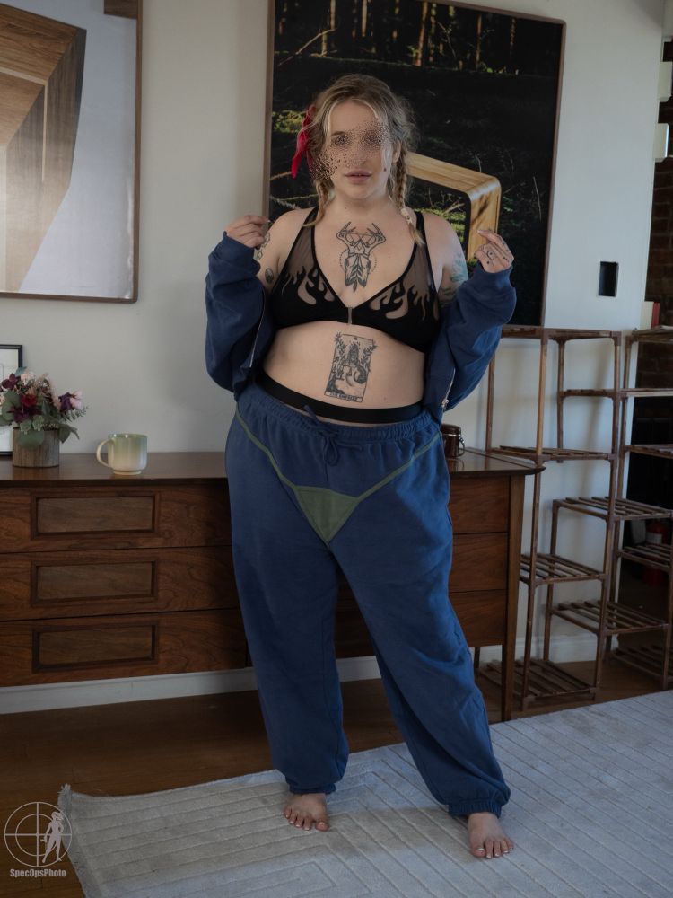 Professional quality photo of Holly Hardwood (BBW Tattooed Woman) standing in front of a dresser. She is wearing a blue tracksuit that has a hilarious green string bikini design on the outside and the top is opened revealing a black lingerie set with an embroidered flame design. Her blonde hair is braided into two cute messy pigtails and there is a hot pink satin bow in her hair. Holly’s face is blurred for privacy.