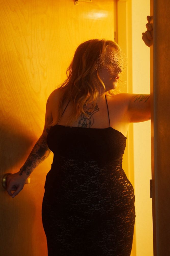 Holly Hardwood (Tall Blonde Tattooed BBW) stepping out of a hotel bathroom. One hand is resting on the doorknob and the other arm is raised and resting on the doorframe. Holly is wearing a form fitting black lace dress and looking over her shoulder back into the bathroom. There is yellow light spilling out of the bathroom lighting the picture and side of her face. 