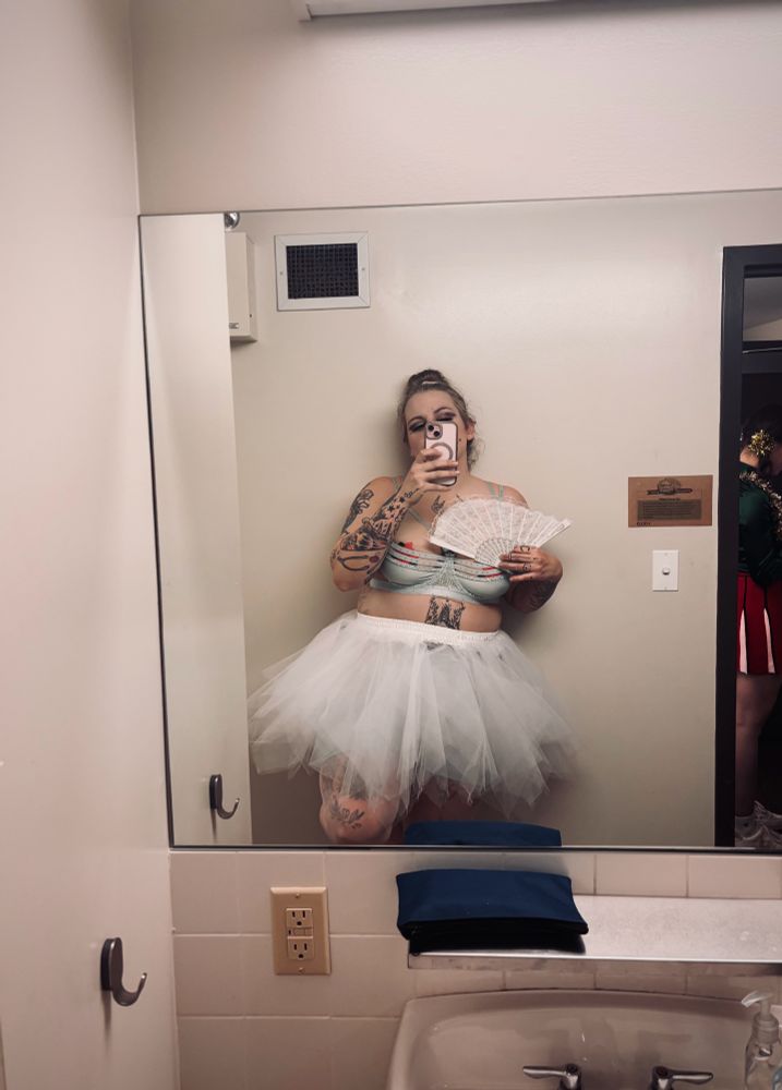 Mirror selfie of Holly Hardwood (Tall Tattooed BBW Woman) leaned back against the wall of a public dressing room. Holly is dressed as a slutty ballerina wearing a powder blue bra, white tutu and ballet pink tights. Her hair is in a high bun and she’s holding a white fan open in front demurely covering her cleavage.