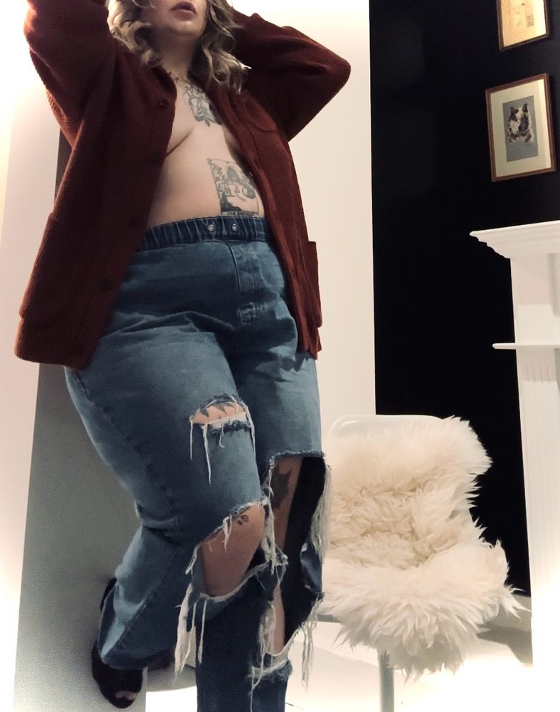 Holly stands leaned against a white pillar with her arms raised to tousle her blonde curled hair. She is wearing torn jeans, stiletto heels and a rust coloured cardigan that is unbuttoned showing her midriff and cleavage. For privacy the top of her face is cropped out just above her juicy luscious lips.