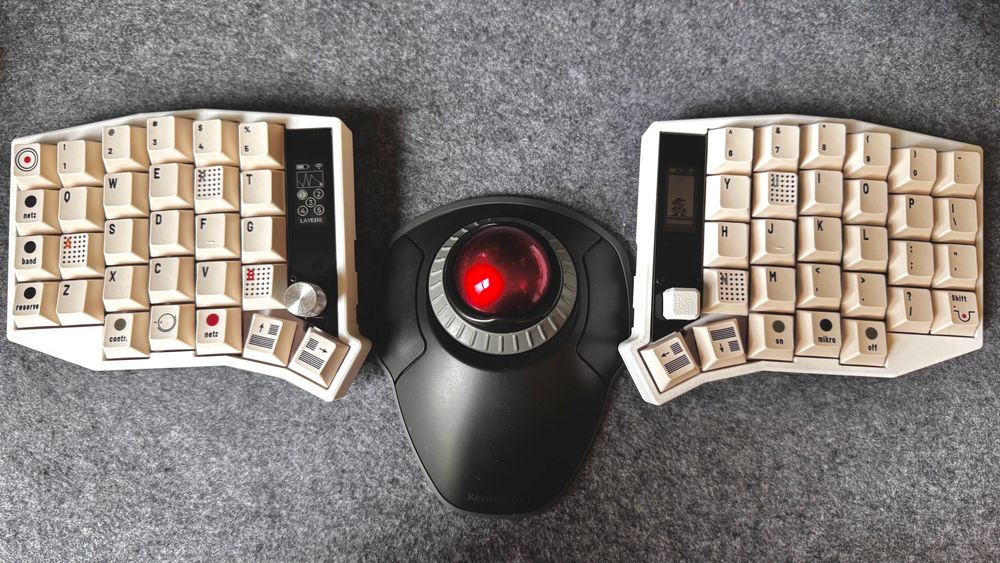 A mechanical split keyboard in an ergonomic, ortho linear format. The keyboard has two screens, a knob, a joystick, and a track ball lives in between the two pieces. The photo is on a gray felt background and the track ball is a Kensington orbit with a red track ball