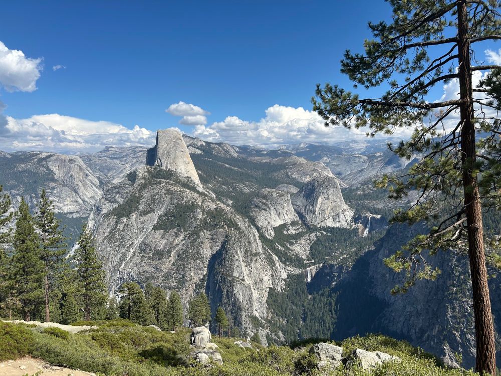 Looking east, from Washburn Point, at the east end of Yosemite Valley, and the Sierra Nevada mountain range. You see huge, dramatic granite expanses, Half Dome, Little Yosemite Valley, Vernal and Nevada falls, Liberty Cap, etc. Scattered throughout are pine trees and olive green shrubs. Above all is a blue sky with fluffy clouds on the horizon.