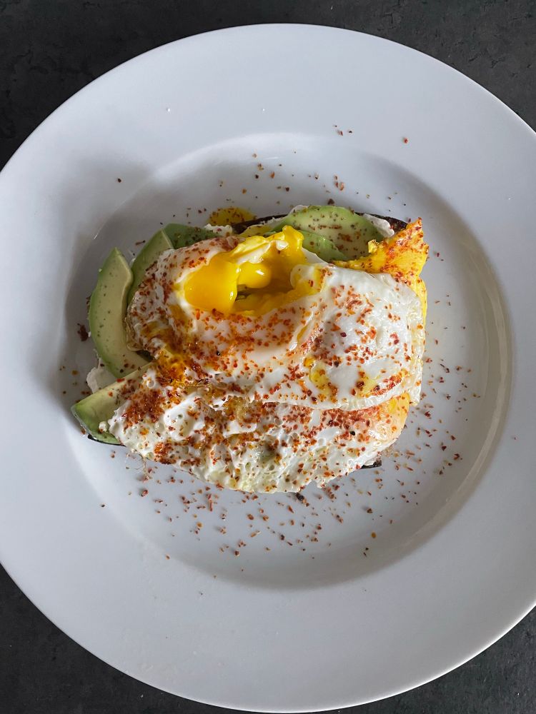 Auf einem weißen Teller liegt eine Scheibe Brot, bestrichen mit Frischkäse, darauf Avocadoscheiben und Spiegelei. Bestreut ist alles mit Chiliflocken.