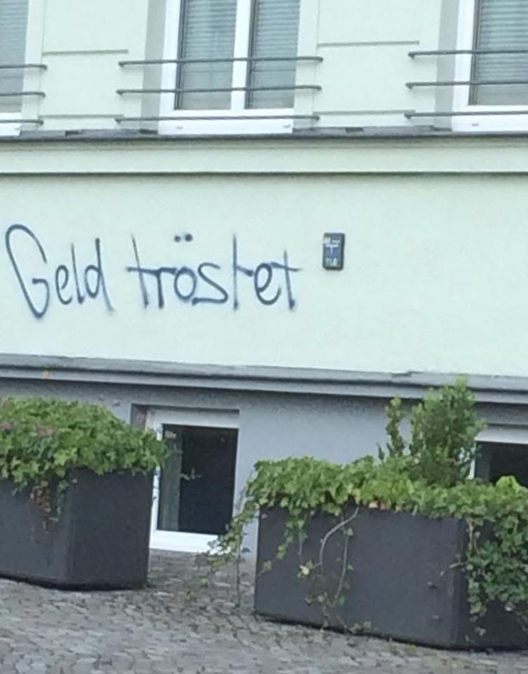 Eine grünliche Hauswand, davor zwei graue Pflanzkübel. An der Wand aufgesprüht „Geld tröstet“