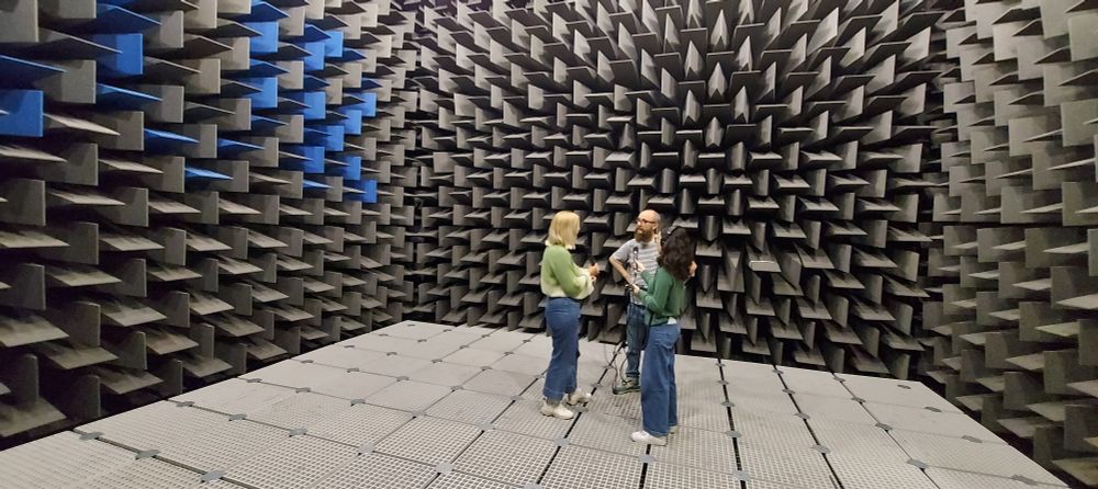 A man and two women with microphones standing in a large room with triangular sound proofing on the walls.