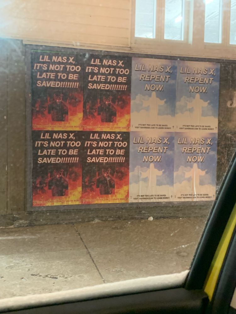 Photo of posters on the side of a wall. There are four posters with fire, a picture of the singer Lil Nas X dressed as the devil, and text that reads “Lil Nas X, it’s not too late to be saved!!!!!!” And four posters with clouds, a white silhouette of Jesus, and text that reads “Lil Nas X, repent now.”