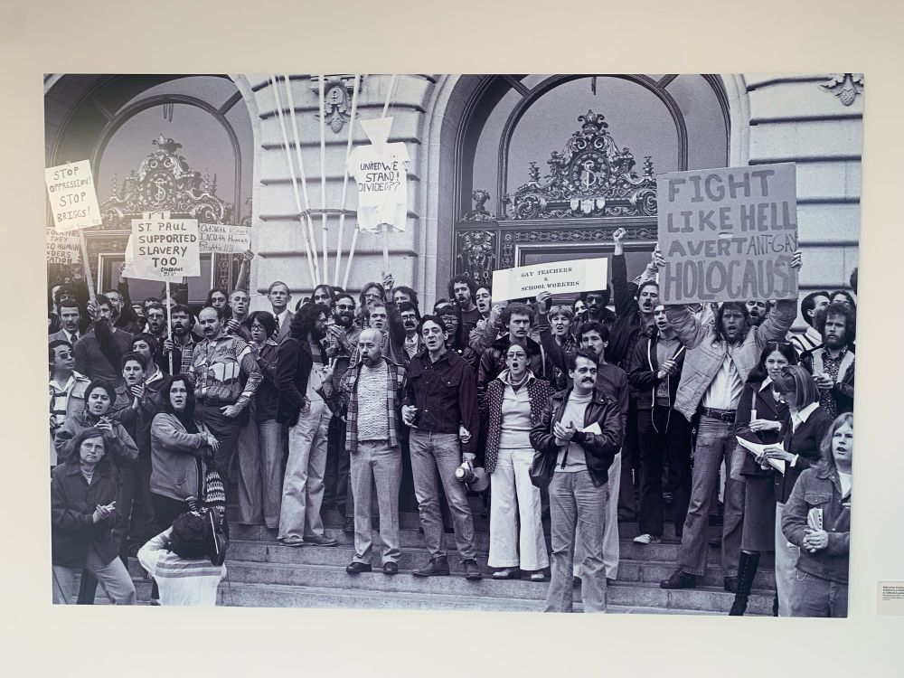 Group of AIDS activists in front of SF City Hall with Harvey Milk. One sign reads Fight Like Hell Avert Our Holocaust.