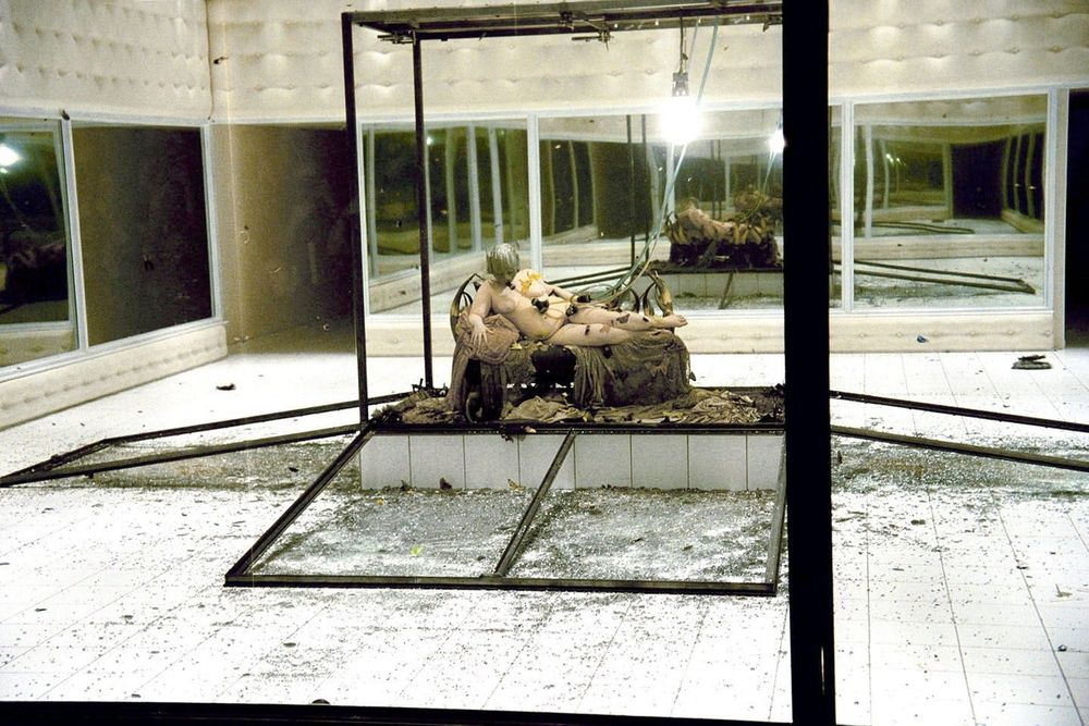 an image from Alexander McQueen's 2001 runway show called Voss. A glass box has been shattered in the middle of a room full of mirrors and harsh lighting. Inside the box a human with a soft belly and thick thighs is lounging on a daybed. They are wearing a full-face metal mask connected to a long hose. They are naked and covered in moths. 