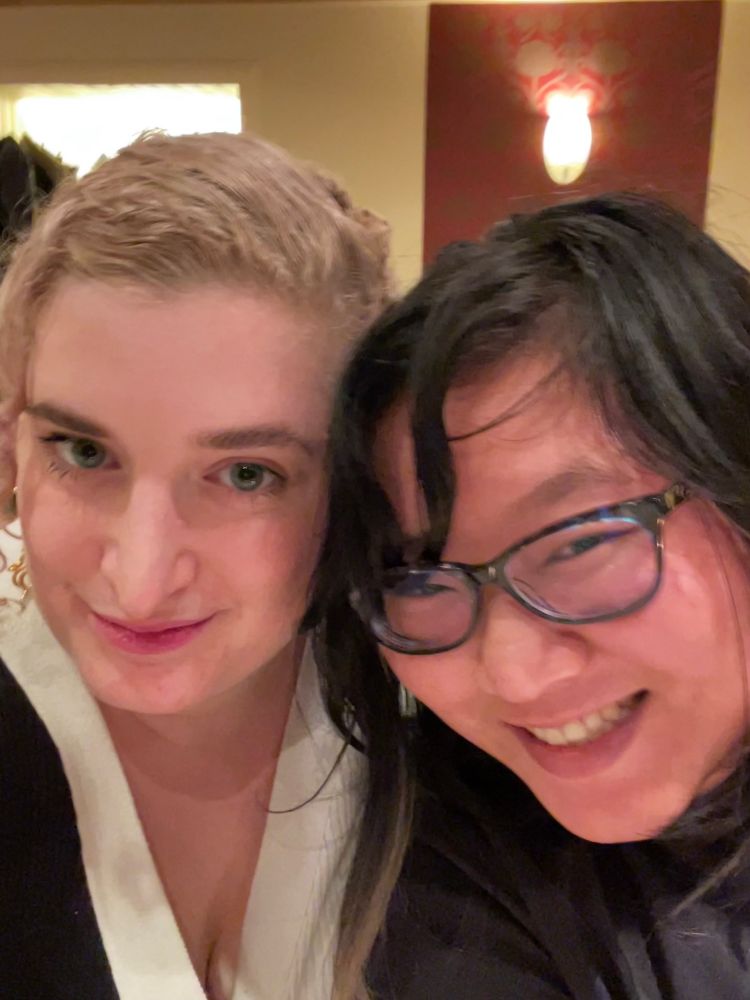 A human with platinum blonde hair and a human with black hair and glasses smile at the camera