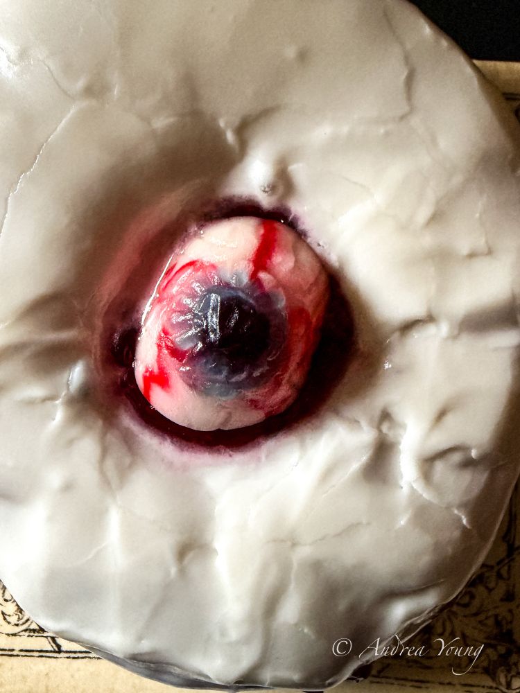 A close-up of a donut with white frosting and a gummy eyeball in the center of it with a blue iris and bright red streaks running through the white parts of the eyeball. 