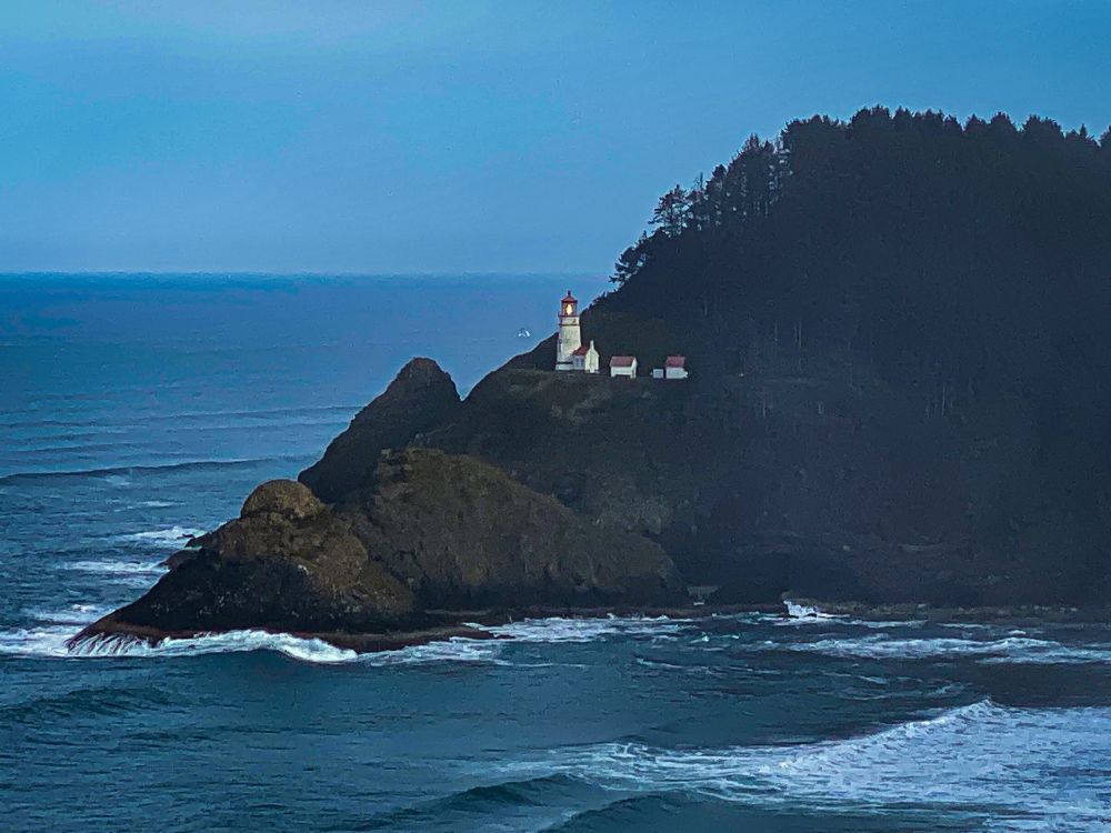 A white lighthouse with red roof-topped buildings at its base. The light house is illuminated and stands up against a forest of trees, on a rocky ledge. A misty sky and ocean are visible in the background, and frothy water is in the foreground.