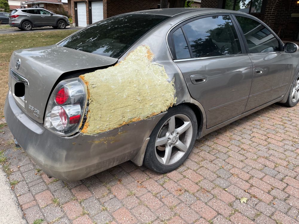 Rear quarter panel of a silver car with a comically bad attempt at auto body repaid. The accordioned panel was filled with domestic spray foam covered with very rough bondo. The tail light is being held in with hidden balled up duct tape behind the light.