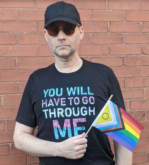 White man with stubble wearing a black baseball hat, sunglasses, and black t-shirt, holding a pride flag, standing in front of a red brick wall. 
