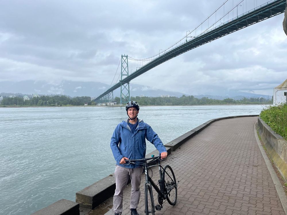 A man wearing grey trousers, a blue jacket and a blue helmet holding his bike near a river with a suspension bridge in the background