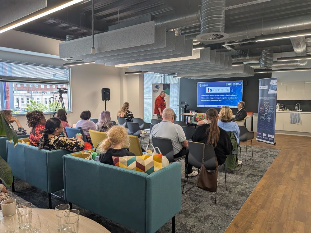 A man in a red shirt giving a presentation with DynAIRx on the screen and an audience facing him listening. 