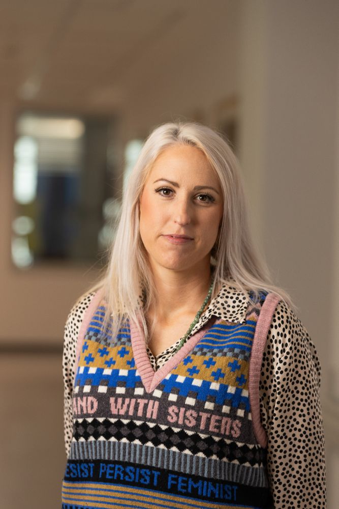 A blond woman wearing colourful clothing looking at the camera. 