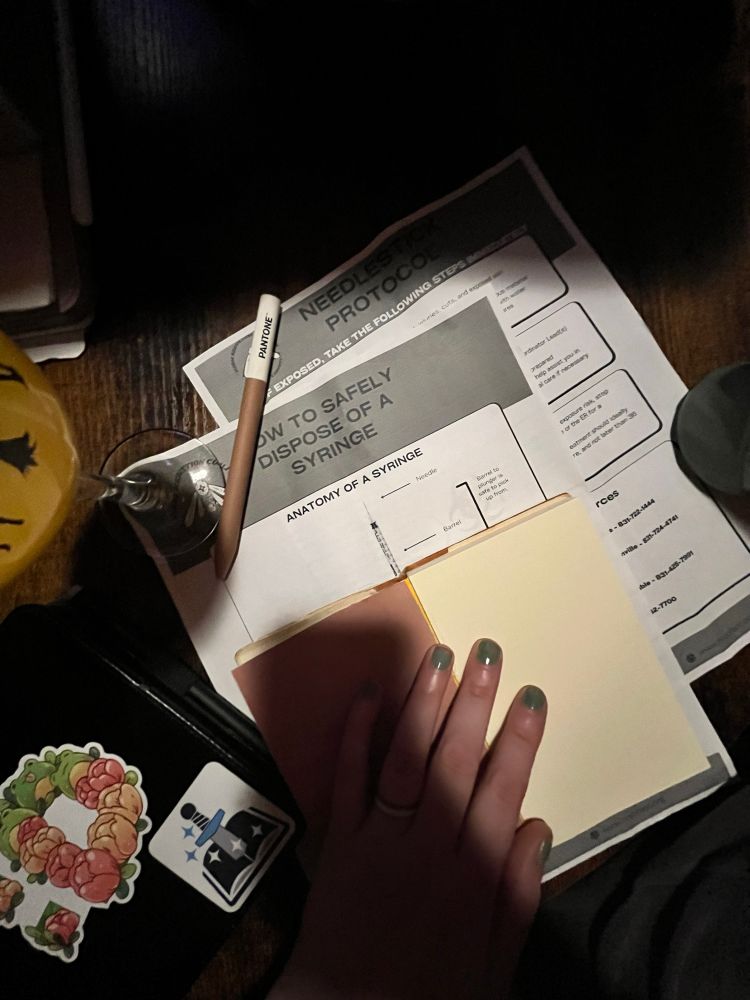 2 documents: NEEDLESTICK PROTOCOL 
& HOW TO SAFELY DISPOSE OF A SYRINGE. A notebook, kobo, and pen on a table. 