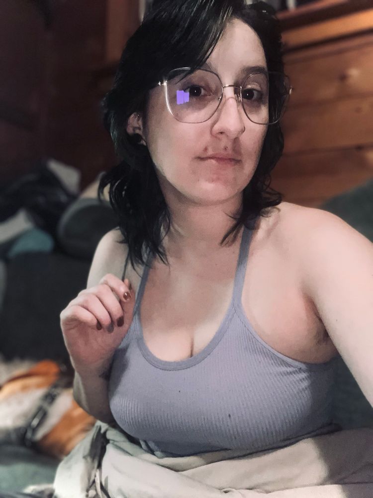 A selfie, I have medium length dark hair, a lavender / gray fitted tank, and large frame glasses on, I’m looking at the camera, my hand up twirling a piece of hair