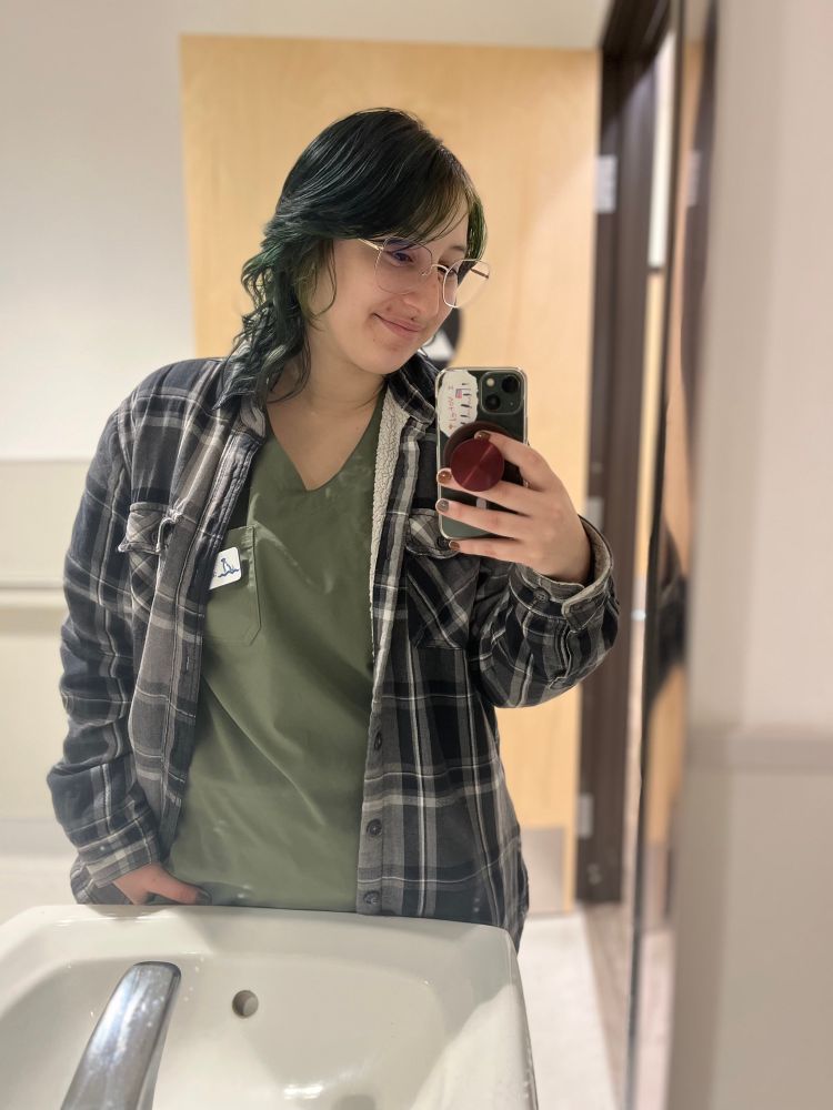 A selfie of me in my work restroom, I have a gray plaid coat on over my green scrubs