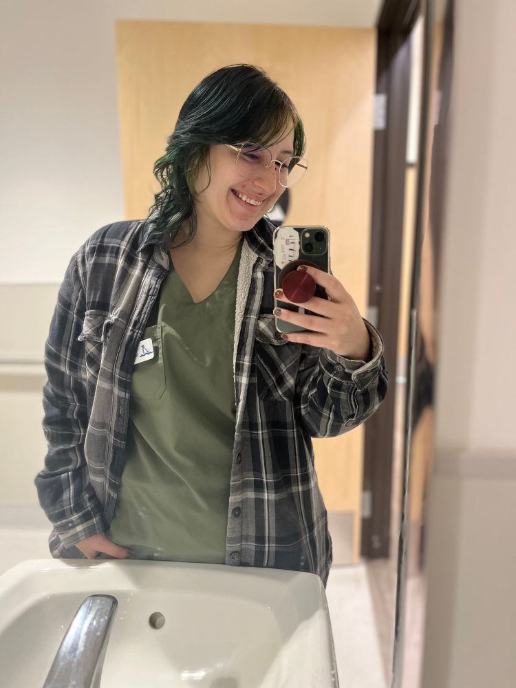 A selfie of me in my work restroom, I have a gray plaid coat on over my green scrubs