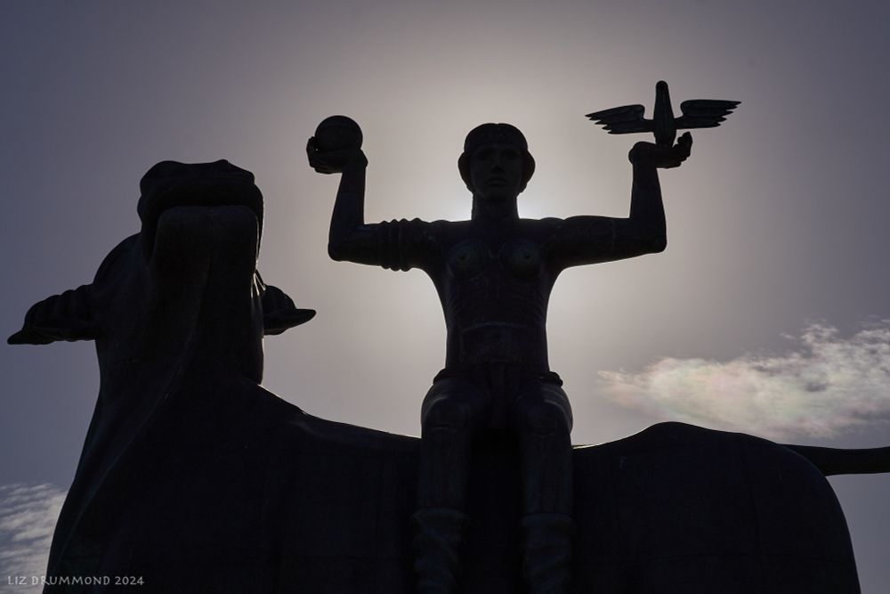 this is a view of the statue of Europa and the bull in silhouette - the sun is behind her body so the photo appears monochrome. On the right hand side of the photo the bull tilts its head up and looks round towards Europa. Europa is sitting completely sideways on the bull's back, very upright and her arms are he'd out sideways at shoulder height and then lower arms and hands are at right angles turned up towards the sky in a pose reminiscent of some of the 'Minoan' statuettes of women that have been discovered. as Europa faces us she holds an orb on the left and a stylised bird on the right. 