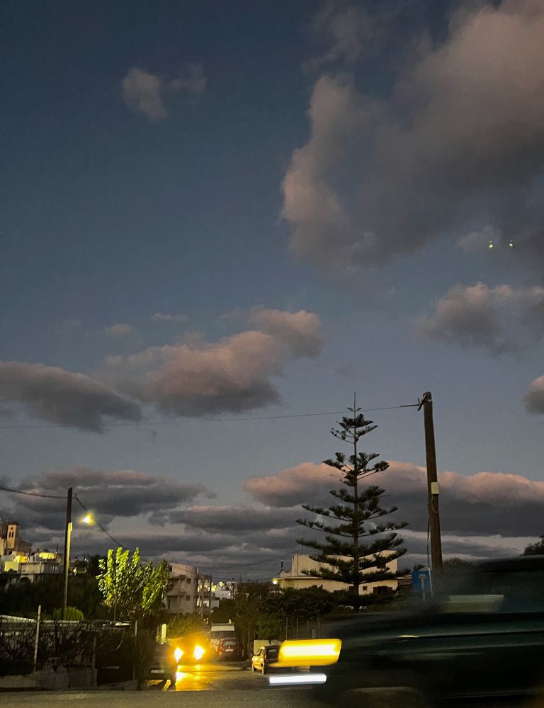Looking down the street from the crossroads into our village in east Crete , it is about 30 minutes after sunset so the sky is fading from blue to dark grey. They are random clouds in the sky in the foreground, lightish grey underneath, lighter on the top and just slightly pink where they are catching the last of the suns rays. In the distance there are banks of clouds that are dark grey underneath and light pink - grey on top.  On the left hand side there is a green mulberry tree catching the light of the streetlight and on the right a rather scraggy conifer is silhouetted against the clouds. A car is coming up the road towards us with its headlights shining and the foreground is partially blurred by a vehicle travelling fast left to right over the crossroads. There are two small pinpricks of white light about 2/3 of the way up on the right hand side which are reflections in the phone camera lens!