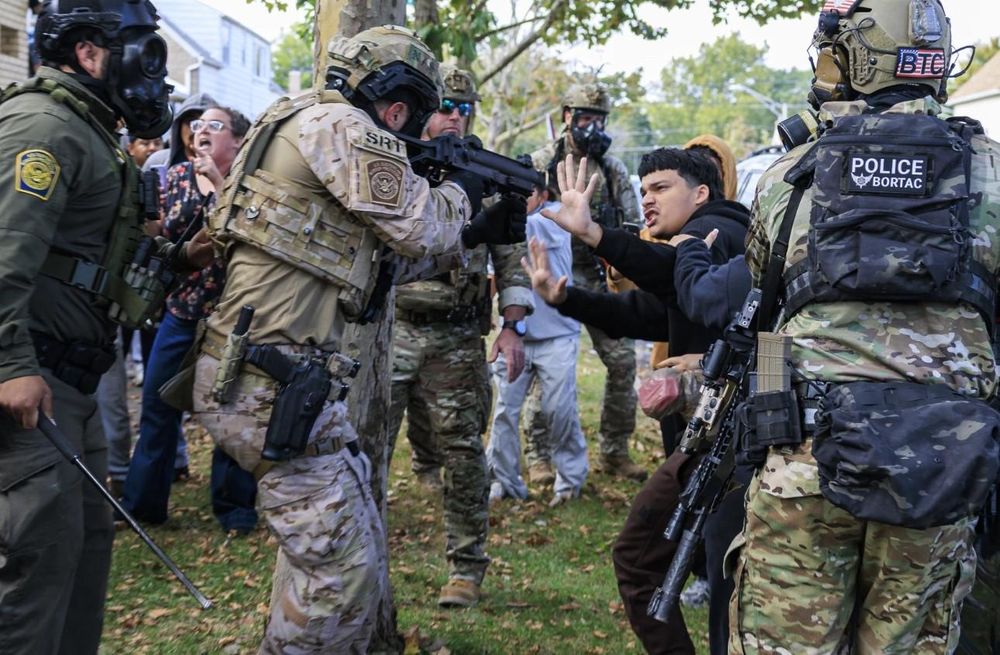 Dressed in full battle-rattle and with his finger on the trigger, a man wearing insignia of a BOTAC team (US Customs agency's special enforcement unit) points a beanbag rifle at the face of a young man who has his hands outstretched to show he's not a threat. The young man appears to be Hispanic and he is surrounded by other BOTAC personnel, all wearing gasmasks and brandishing firearms, plus a CBP agent in the green Border Patrol uniform with a gas mask who is brandishing a riot whip. In the background there is a middle-aged white lady who appears to be trying to argue the CBP should let her past so she can help the young man. Another CBP man in a gas mask is holding another Hispanic youth against a tree by the collar of their white tshirt. Photo by Chicago Sun Times.