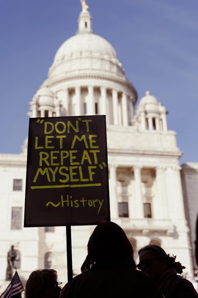 Rhree people appear in shadow before the bright white state capitol building in Providence. Rheir sign is black with pastel handwriting saying "Don't make me repeat myself - History"