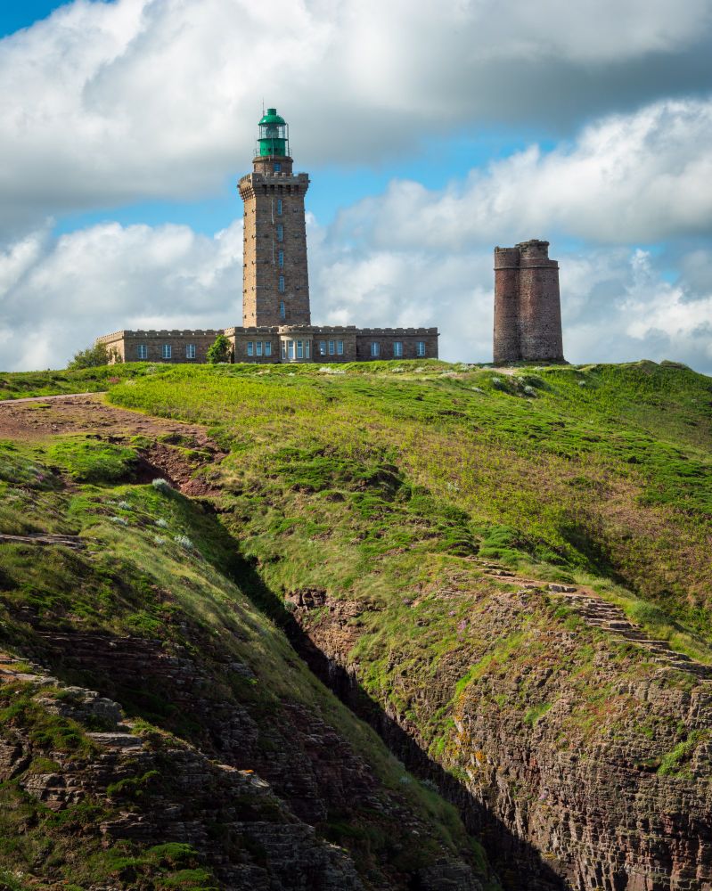 Ein imposanter Leuchtturm aus Stein mit einem grünen Laternenraum steht auf einer Klippe mit grünen Wiesen. Rechts daneben befindet sich eine alte, runde Ruine eines früheren Turms. Die Klippen sind von rötlich-braunem Gestein durchzogen, mit tiefen Furchen und grasbewachsenen Flächen. Der Himmel ist teils bewölkt, mit weißen Wolken vor einem blauen Hintergrund.