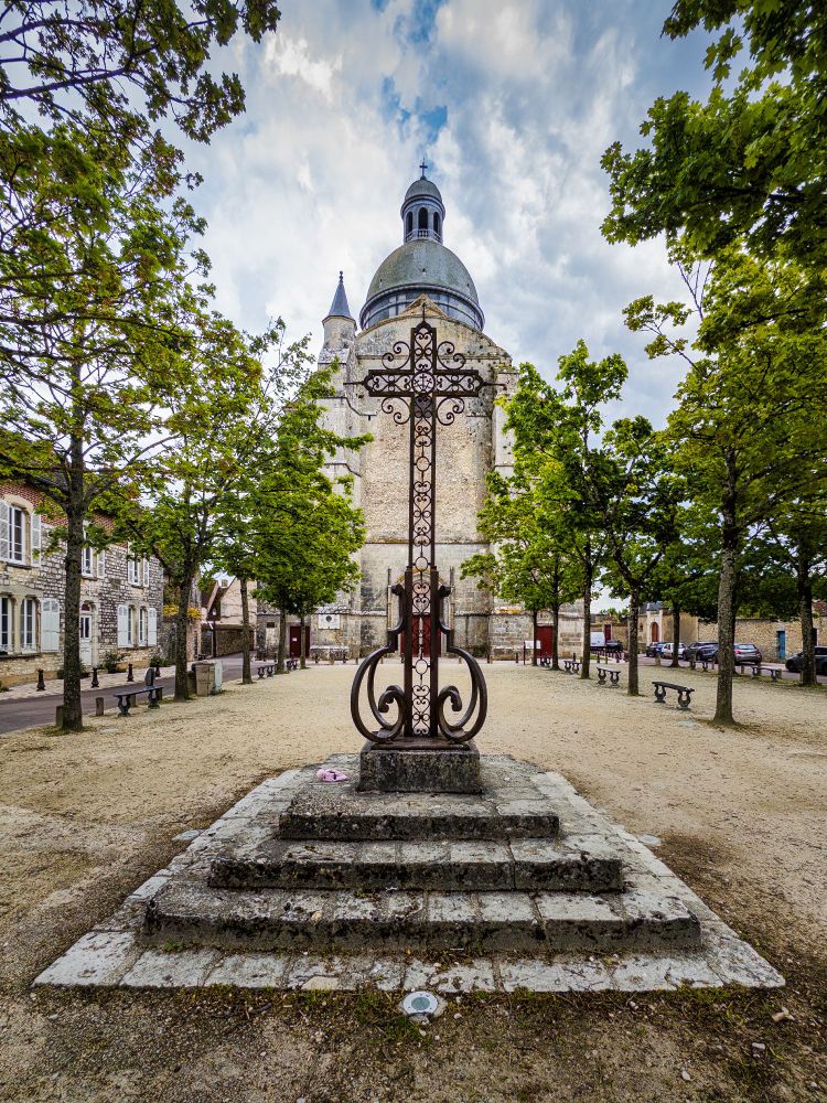 Historisches Kreuz auf einem mehrstufigen Sockel, verziert mit filigranen Eisenornamenten. Im Hintergrund erhebt sich eine imposante Kirche mit großer Kuppel und kleinerem Turmaufsatz. Der Platz ist von Bäumen gesäumt und mit Sitzbänken ausgestattet, flankiert von alten Gebäuden und einer Straße mit parkenden Autos. Der Himmel zeigt eine Mischung aus Wolken und blauen Lücken.