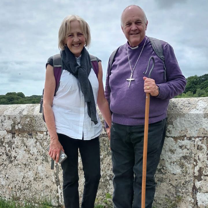 This photograph shows two people stood by a white stone wall. The sky is cloudy and the tree tops are peering up over the top of the wall