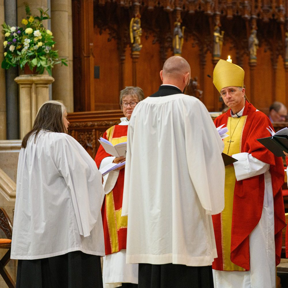 Image shows the 2024 Ordination Service in the cathedral. Two people dressed in white stood in front of two people in red and yellow. They are holding white booklets. There are some flowers to the top left.