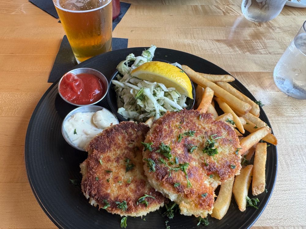Two Dungeness crabcakes on a bed of fries with slaw, tartar sauce, a slice of lemon, ketchup, and a beer in the background