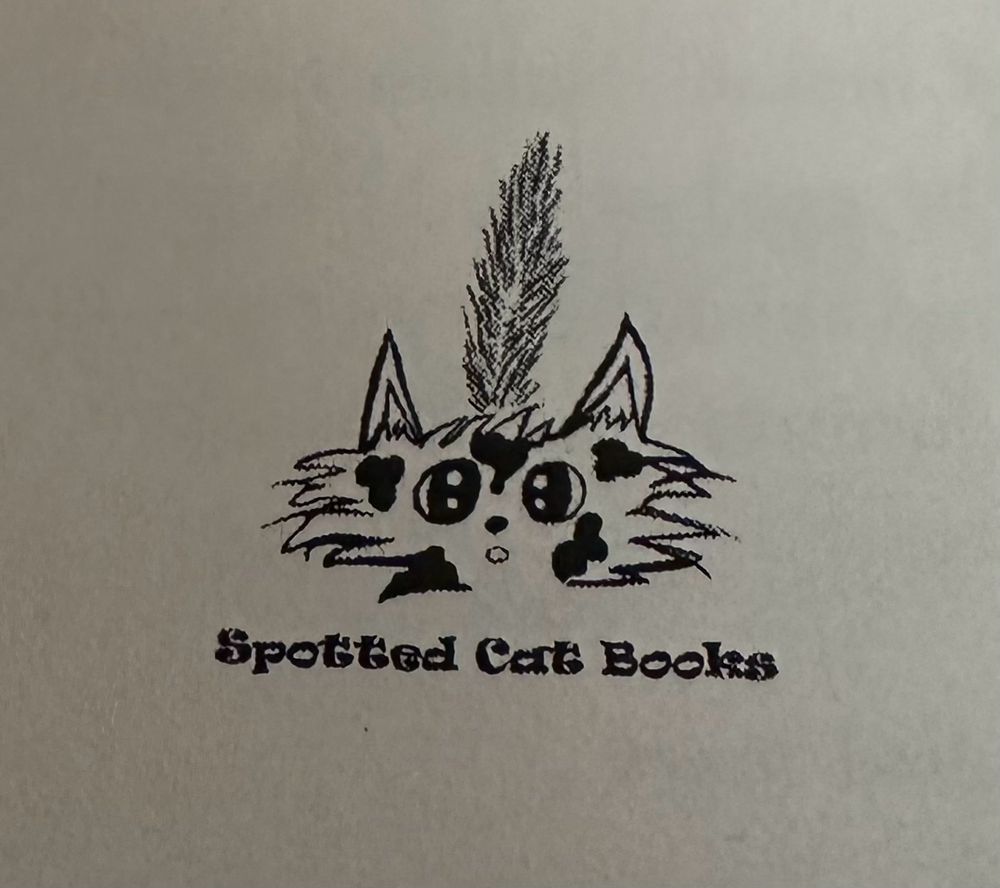 Logo of a silly and slightly startled looking fuzzy white cat with a bushy tail sticking up behind him. He’s got black spots on his face and big round eyes that are slightly cross eyed. Underneath him is Spotted Cat Books in a rounded typeface.