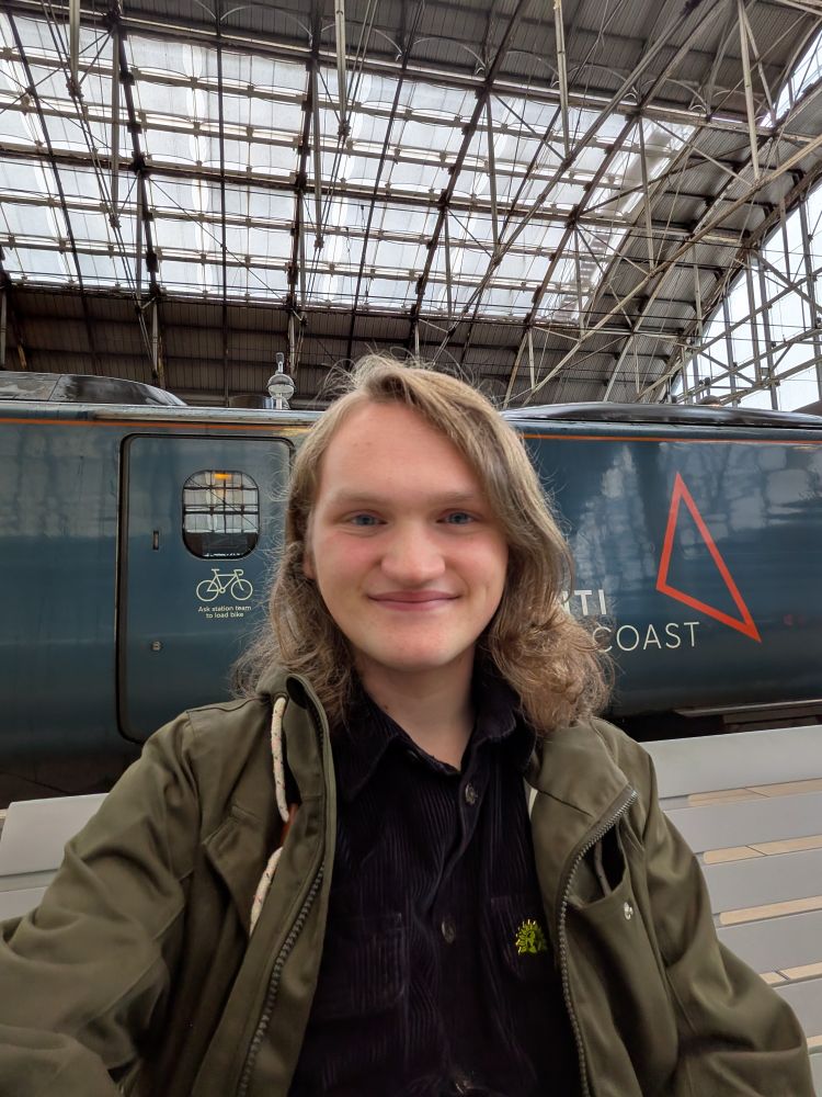 A photo of Finn White. They are wearing a black shirt and a green coat. Behind them is a train.