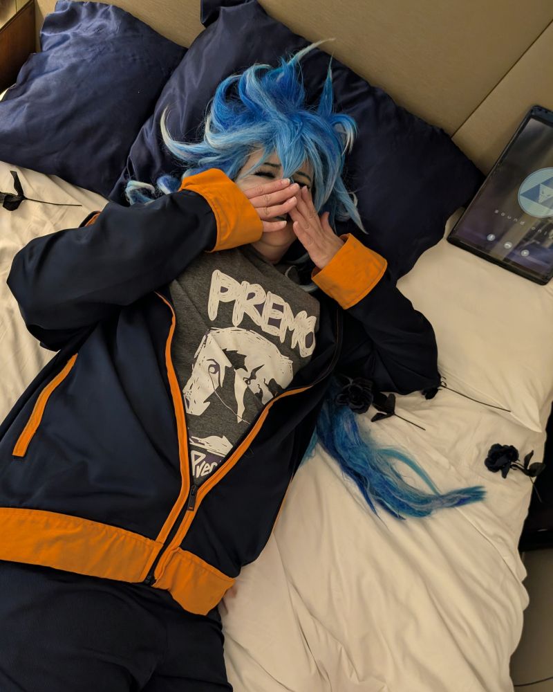 A photograph picture of a cosplayer in the loungewear outfit of the character Idia Shroud from Disney Twisted Wonderland mobile game, laying on a bed with black roses and a tablet.