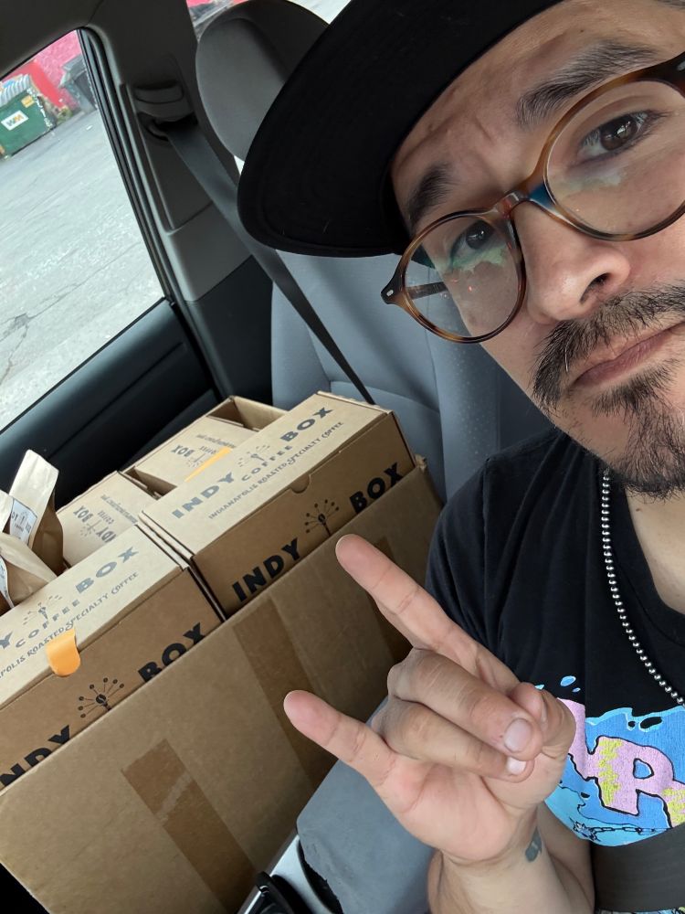 Me, in the drivers seat of my car, at a stop light, waving at the camera. In the passenger's seat is a large cardboard box filled with boxes that say “Indy Coffee Box.”