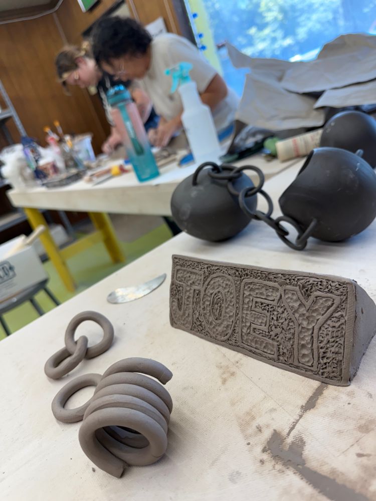 A ceramic studio interior, two adult students in the background, slightly blurred, stand at a studio table. In focus is a clay name tag for “JOEY” with textured letters, and a coil of clay used to make rings for a ceramic chain.